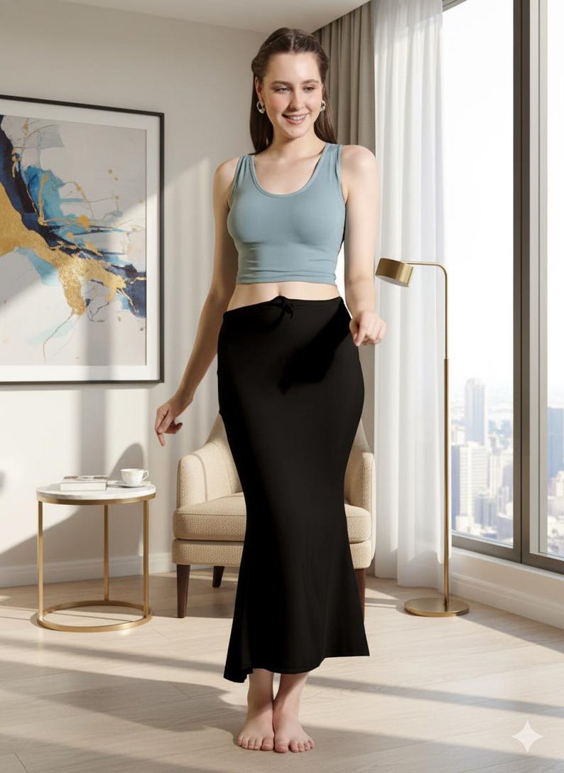 May include: A woman wearing a light blue tank top and a black, floor-length skirt. The skirt has a slight flare at the bottom. The woman is standing in a well-lit room with a window and a gold lamp.