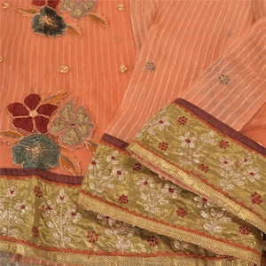 May include: A peach-colored fabric with vertical white stripes, featuring large embroidered floral appliqués in dark red and muted green. The hem is finished with a wide, metallic gold border decorated with small red and white floral embroidery.