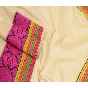 May include: A cream-colored fabric featuring a wide border with a vibrant pink floral pattern, followed by horizontal stripes in yellow, orange, and green. The textile has a smooth, woven texture with visible folds and a consistent color palette.
