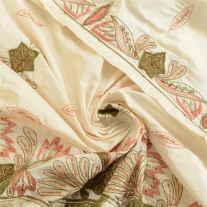 May include: An off-white fabric featuring intricate embroidery and beadwork in shades of coral pink and olive green. The design includes stylized floral and leaf motifs arranged in a border pattern, with small scattered leaf shapes across the surface.
