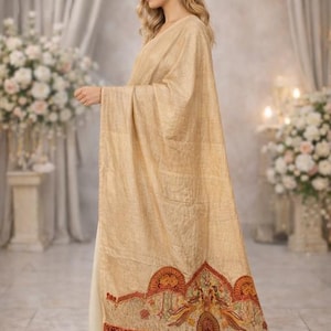 May include: A long, beige shawl with a textured finish, draped over a light-colored garment. The bottom edge features a wide, intricate border with red, orange, and gold embroidery depicting stylized floral and bird motifs in a repeating pattern.
