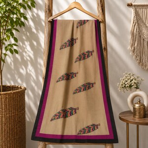 May include: A rectangular beige scarf featuring a repeating pattern of embroidered feathers in shades of green, purple, and orange. The scarf is framed by a vibrant magenta border and a thin black outer edge, hanging from a wooden hanger.