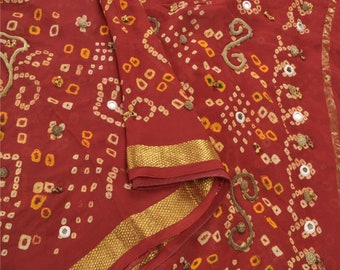 May include: A deep red fabric featuring a traditional tie-dye pattern with small yellow and white square motifs. The textile is embellished with circular mirrors and gold-toned metallic embroidery, finished with a wide gold textured border.