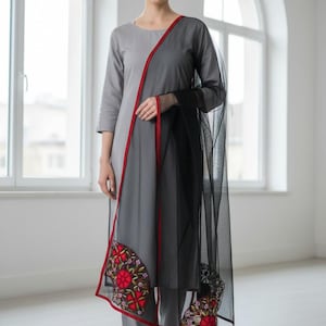 May include: A grey kurta suit with a black dupatta. The kurta has a red trim and embroidered floral designs. The black dupatta has a sheer, net-like texture. The outfit is worn with grey trousers and sandals.