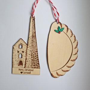 May include: Two wooden Christmas decorations. One is a brown and white silhouette of a building with the text "Merry Christmas! Cornwall" and a red heart. The other is a brown and gold silhouette of a pastry with a green holly sprig.
