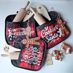May include: A festive Christmas baking set featuring red and black plaid oven mitts with seasonal text and gingerbread decorations. Wooden rolling pins and spoons are included, alongside a decorative bow.