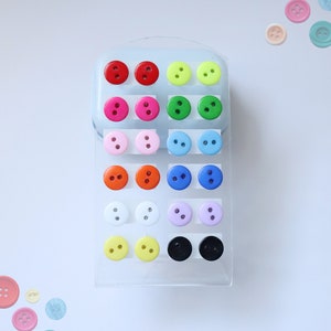 May include: A set of 18 colourful button earrings displayed on a clear plastic card. The earrings are arranged in rows of two, with each row featuring a different colour. The colours include red, pink, green, blue, orange, white, yellow, black, and purple.