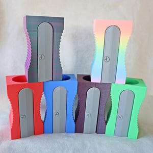 May include: A selection of large, brightly coloured pencil sharpeners. The range includes red, blue, green, purple, and a rainbow gradient. Each sharpener has a grey blade and a cross-shaped detail. The sharpeners are stacked, highlighting their distinctive design.