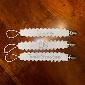 May include: Three white fabric baby bib clips with scalloped edges. The top clip says "Turner", the middle clip says "TJS", and the bottom clip says "Turner Stevenson".