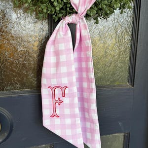 May include: A pink and white gingham ribbon with a red embroidered letter "F" hangs from a green wreath. The ribbon is tied in a knot, creating a decorative accent. The background is a dark blue door.