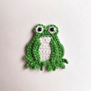 May include: A handmade crochet frog applique in shades of green and white. The frog has large, round eyes with black pupils. The body is a vibrant green, with a white belly. This whimsical applique is perfect for crafting projects.