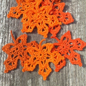 May include: Several handmade, orange crochet maple leaves are displayed on a weathered wooden surface. The crochet work is detailed, with openwork patterns. The leaves are in various shades of orange, creating a textured appearance. A seasonal, autumnal theme.
