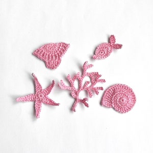 May include: Five pink crocheted sea-themed decorations: a starfish, a seashell, a fish, and two pieces of coral.