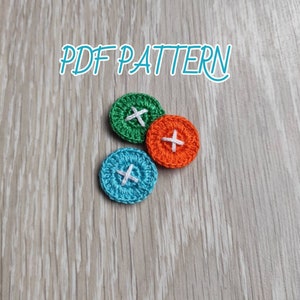 May include: Three crocheted buttons with white stitching in the shape of an 'X' on a woodgrain background. The buttons are green, orange, and blue.