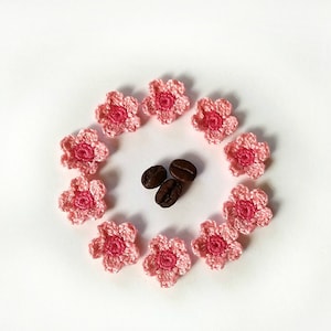 May include: A circle of pink crocheted flowers surrounds three coffee beans on a white background.