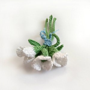 以下が含まれることがあります： 白いベル型の花、緑の葉と茎で作られた手作りのかぎ針編みのブーケ。茎は水色の糸で結ばれています。花は小さくコンパクトなクラスターに配置されており、装飾的なアクセントに最適です。