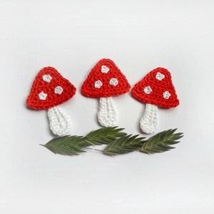May include: Three crocheted red and white mushrooms with white stems. The mushrooms are arranged in a row on a white background with green leaves.