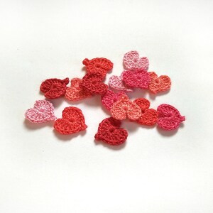 Puede incluir: Una colección de corazones tejidos a crochet en varios tonos de rosa y rojo. Los corazones están dispuestos en un grupo sobre un fondo blanco.