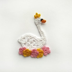 May include: A white crocheted swan wearing a yellow crown, with an orange beak and black eye. The swan is encircled by pink and yellow crocheted flowers. The craft item is on a white background.
