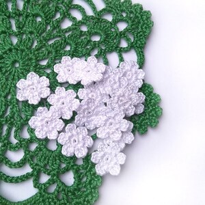 May include: A close-up of a green crocheted doily with a floral design. Several small, white crocheted flowers are scattered on the doily. The doily and flowers are on a white background. The handmade items are likely for home decor.