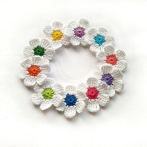May include: A circular arrangement of crocheted white daisy flowers with colourful centres. The flower centres are yellow, purple, teal, orange, green, blue, pink, and red. The daisies are arranged in a circle on a white background.