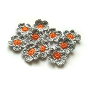 May include: A cluster of small, crocheted flowers. Each flower features grey petals and a vibrant orange centre. The flowers are arranged on a white surface, giving a handmade and cheerful appearance.