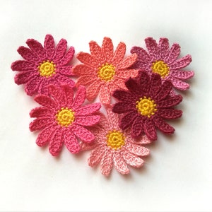 May include: A collection of six crocheted daisy flowers in shades of pink, coral, and burgundy, arranged in a heart shape. Each flower has a yellow center and detailed petal work, set against a white background.