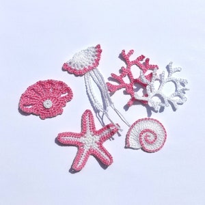 Può includere: Un set di cinque decorazioni all'uncinetto a tema marino in rosa e bianco. Il set comprende una stella marina, una conchiglia, una medusa e due pezzi di corallo.