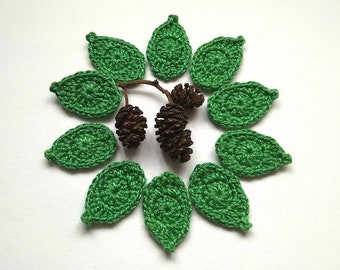 May include: A collection of handmade crochet leaves in a vibrant green arranged in a circular pattern. Two small brown pinecones and a twig are in the center. The leaves are detailed with visible stitch work, suggesting a craft project.