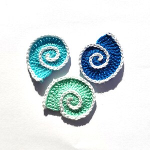 May include: Three crocheted seashell appliques in shades of blue and green, each with a white spiral centre and white edging. The appliques are arranged on a white background, showcasing the handmade details and vibrant colours.
