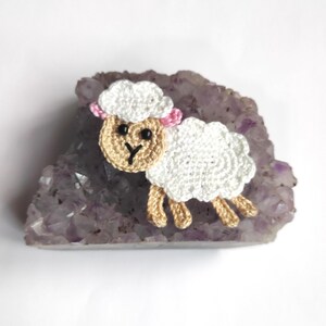 May include: A crocheted sheep with a white body, beige face and legs, and a white fluffy head with pink ears. The sheep is resting on a large, rough-edged, purple amethyst geode. The background is white.