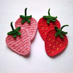 May include: Four handmade crochet strawberry coasters. Two are light pink, and two are red. Each coaster has a green stem and leaves. Yellow stitches represent the seeds. These coasters are ideal for adding a touch of whimsy to your home decor.