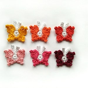 May include: Six handmade crochet butterfly appliques in various colors: yellow, orange, red, pink, fuchsia, and burgundy. Each butterfly has white accents and is displayed on a white background. These appliques are ideal for crafting projects.