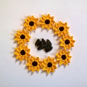 May include: A circular arrangement of handmade crochet sunflowers with yellow petals and dark brown centers. A small pile of black sunflower seeds sits in the center of the floral design. The flowers are set against a white background.