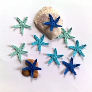 May include: A collection of handmade crochet starfish in shades of blue and green, arranged with a seashell and small stones. The starfish vary in color from light teal to deep blue. The image is set against a white background.