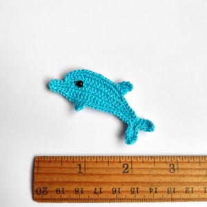 May include: A small, turquoise crochet dolphin with a black eye is displayed on a white background. The dolphin is approximately 2 inches long. A wooden ruler is placed below the dolphin for size reference. The dolphin is a handmade craft item.