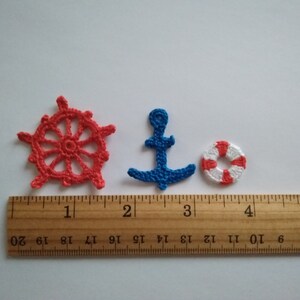 May include: Three crocheted nautical themed ornaments: a red ship's wheel, a blue anchor, and a red and white life preserver. The ornaments are displayed on a wooden ruler with inch markings.