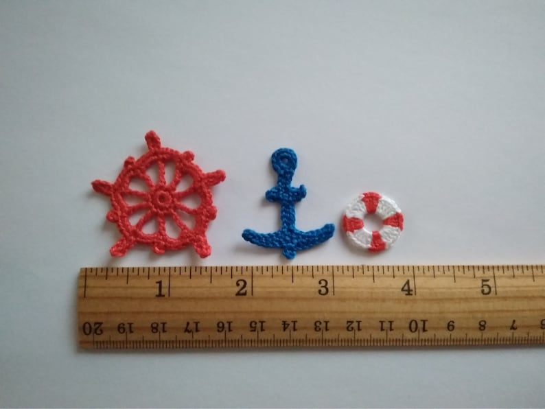 May include: Three crocheted nautical themed ornaments: a red ship's wheel, a blue anchor, and a red and white life preserver. The ornaments are displayed on a wooden ruler with inch markings.