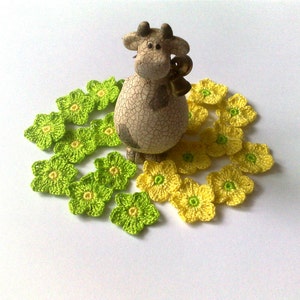 May include: A ceramic cow figurine with a bell around its neck, surrounded by crocheted flowers. The flowers are in two colors: light green and yellow, with a yellow center. The figurine has a cracked, textured appearance.