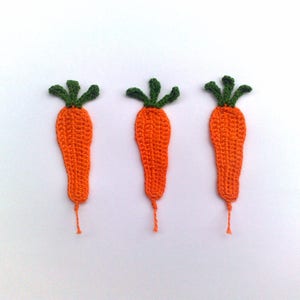 May include: Three handmade crochet carrots with orange bodies and green tops are arranged on a white background. Each carrot has a small orange thread extending from the bottom. The carrots are a whimsical, handcrafted item.