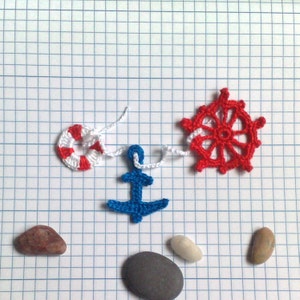 May include: A blue anchor, a red and white life preserver, and a red crochet steering wheel are strung together with white yarn. There are three gray stones and one brown stone on a white grid background.