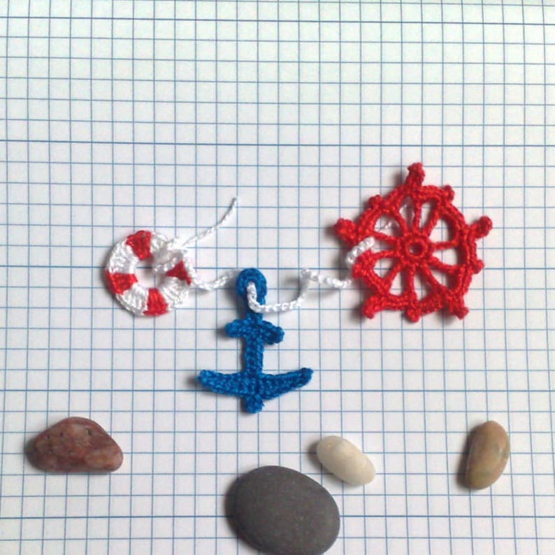 May include: A blue anchor, a red and white life preserver, and a red crochet steering wheel are strung together with white yarn. There are three gray stones and one brown stone on a white grid background.