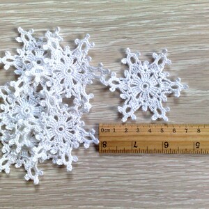 以下が含まれることがあります： 木製の表面に、さまざまなサイズの白いかぎ針編みの雪の結晶がいくつか表示されています。定規は、雪の結晶の直径が約7.6cmから10.2cmであることを示しています。雪の結晶は、冬の手工芸品に最適な、複雑な透かし彫りのデザインが特徴です。
