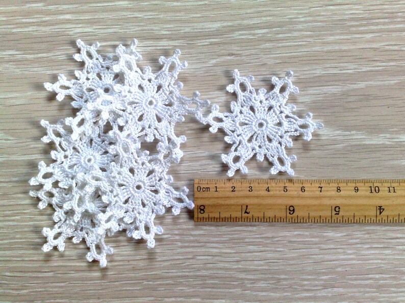 以下が含まれることがあります： 木製の表面に、さまざまなサイズの白いかぎ針編みの雪の結晶がいくつか表示されています。定規は、雪の結晶の直径が約7.6cmから10.2cmであることを示しています。雪の結晶は、冬の手工芸品に最適な、複雑な透かし彫りのデザインが特徴です。
