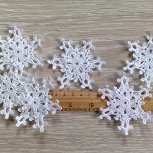 以下が含まれることがあります： さまざまなサイズの白いかぎ針編みの雪の結晶が6つ、明るい木製の表面に配置されています。 各雪の結晶は、中央の円と放射状のポイントを備えた、詳細なオープンワークデザインです。 定規が近くに置かれています。