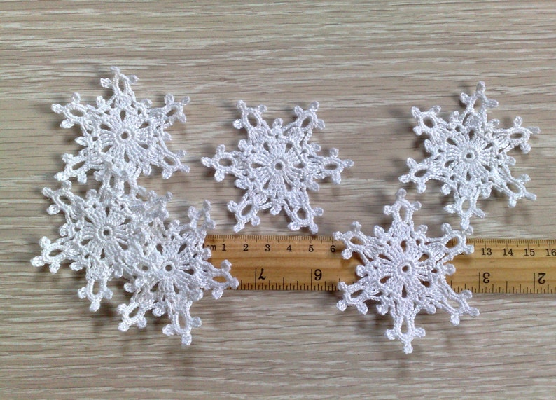 以下が含まれることがあります： さまざまなサイズの白いかぎ針編みの雪の結晶が6つ、明るい木製の表面に配置されています。 各雪の結晶は、中央の円と放射状のポイントを備えた、詳細なオープンワークデザインです。 定規が近くに置かれています。