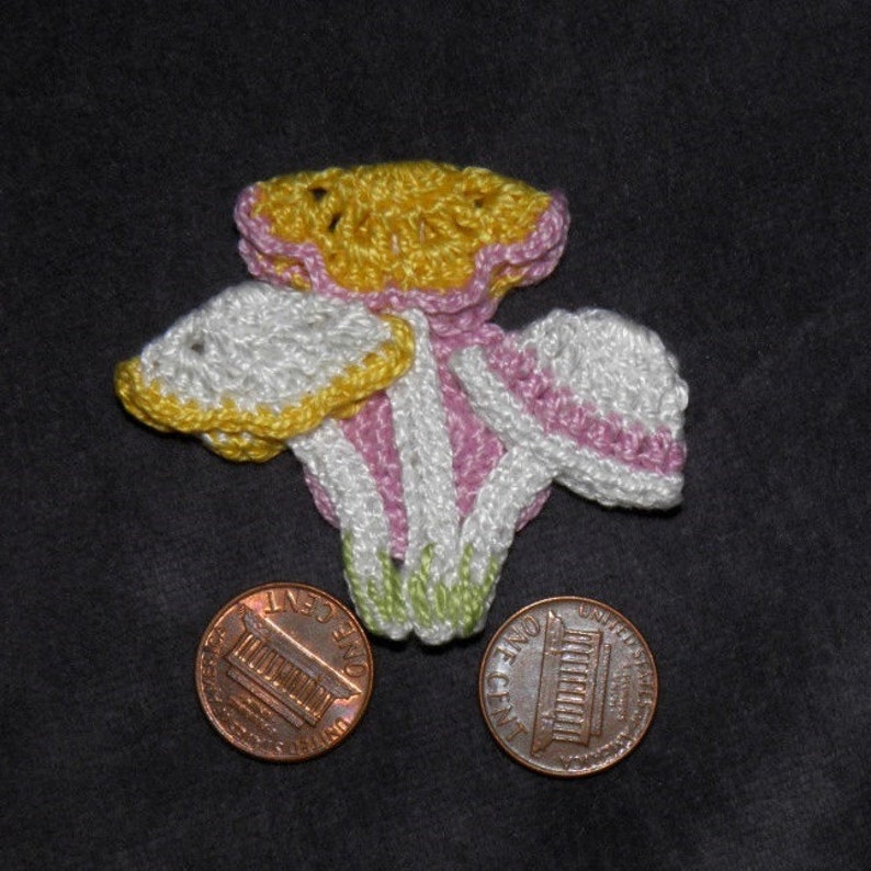 Puede incluir: Una flor de ganchillo hecha a mano en amarillo, blanco y rosa, con detalles verdes. Dos monedas de un centavo se colocan debajo de la flor para comparar el tama&ntilde;o. La flor tiene un dise&ntilde;o en capas sobre un fondo gris oscuro.