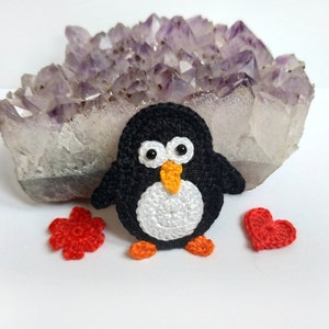 May include: A handmade crochet penguin with a white belly, black body, orange feet and beak, and white eyes. A red crochet flower and heart are next to the penguin. A large amethyst crystal cluster is in the background.
