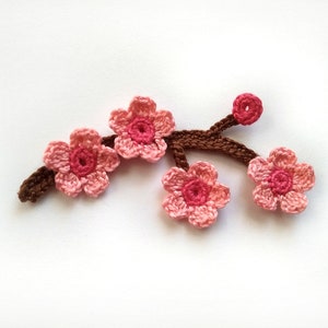 May include: A crocheted floral branch with four pink flowers, each with a darker pink centre, and a small bud. The brown branch supports the flowers. The craftwork is set against a white background.