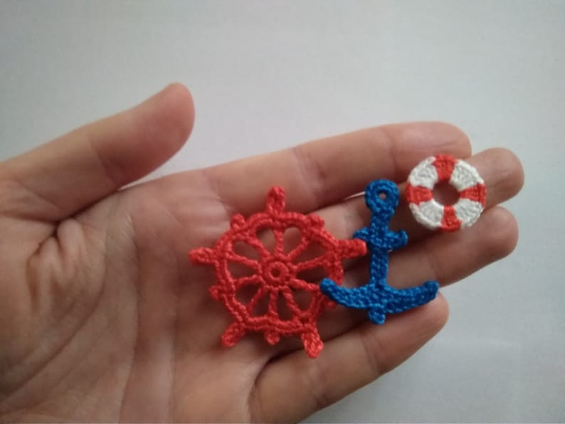 May include: A set of three crocheted nautical themed ornaments. The ornaments are a red and white life preserver, a blue anchor, and a red ship's wheel.